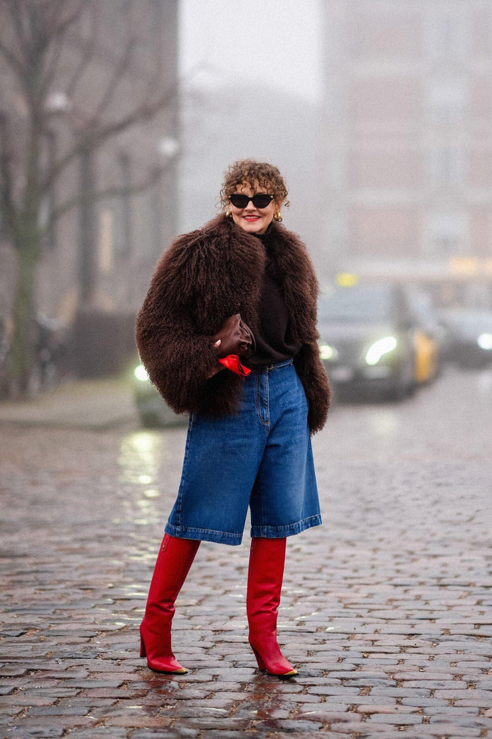 Renia Jaz wears sunglasses, golden earrings, red lipstick, a brown oversized fluffy faux fur jacket, a leather bag, cropped blue denim jeans pants, red boots with high heels, outside Rolf Ekroth, during the Copenhagen Fashion Week AW24, 2024 in Copenhagen, Denmark. Photo by Edward Berthelot/Getty Images